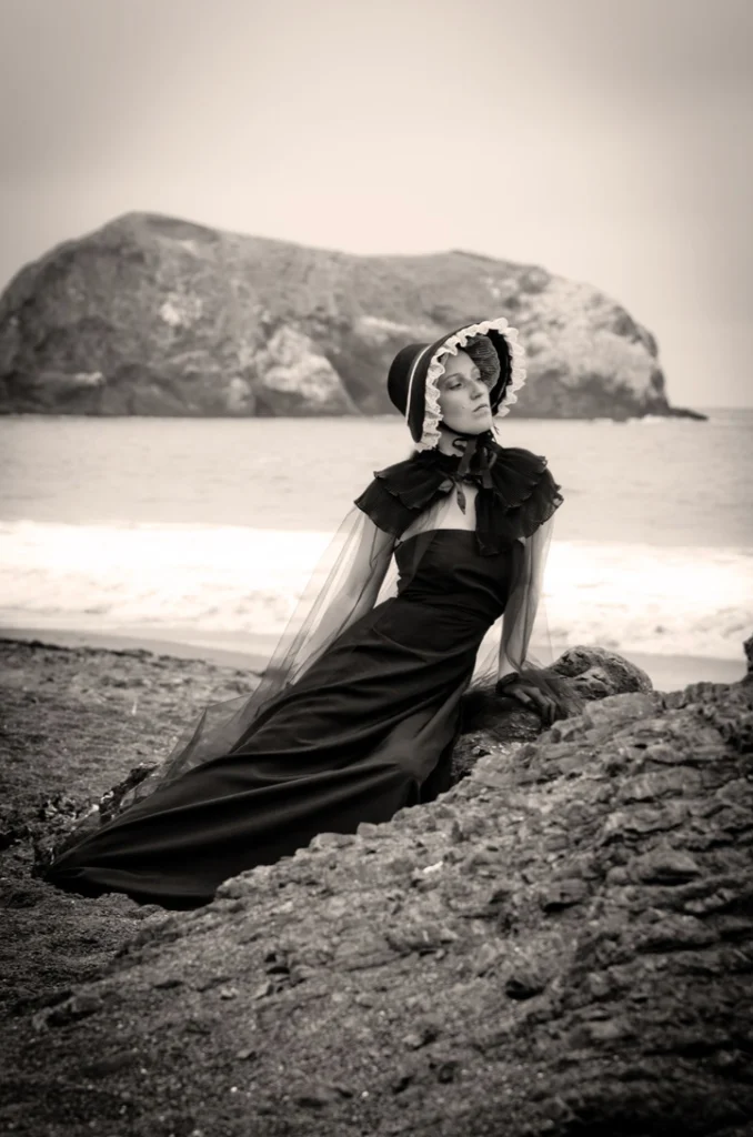 Femme en robe noire et capeline allongée sur les rochers face à la mer