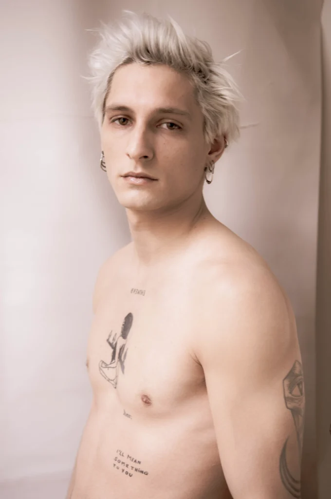 Jeune homme aux cheveux platine, torse nu et tatoué, de trois quarts